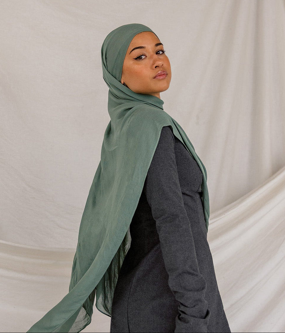 Person wearing a green hijab, dark top, and gray pants against a beige draped curtain background