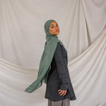 Person wearing a green hijab, dark top, and gray pants against a beige draped curtain background