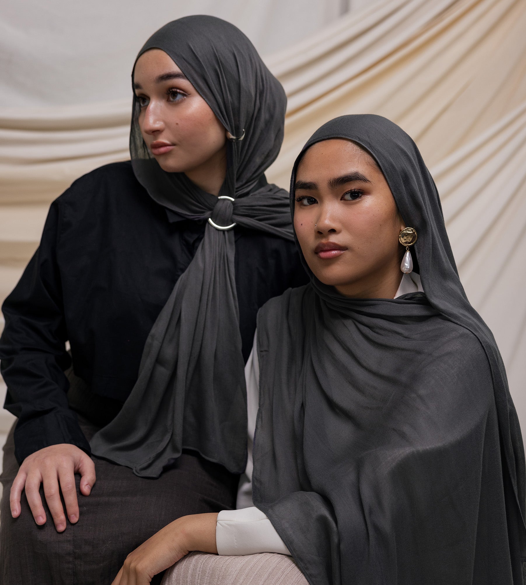 Two women wearing dark gray modal hijabs, one seated and one standing, posing with neutral expressions.