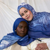 Two women wearing blue with white shell printed hijabs against a white background