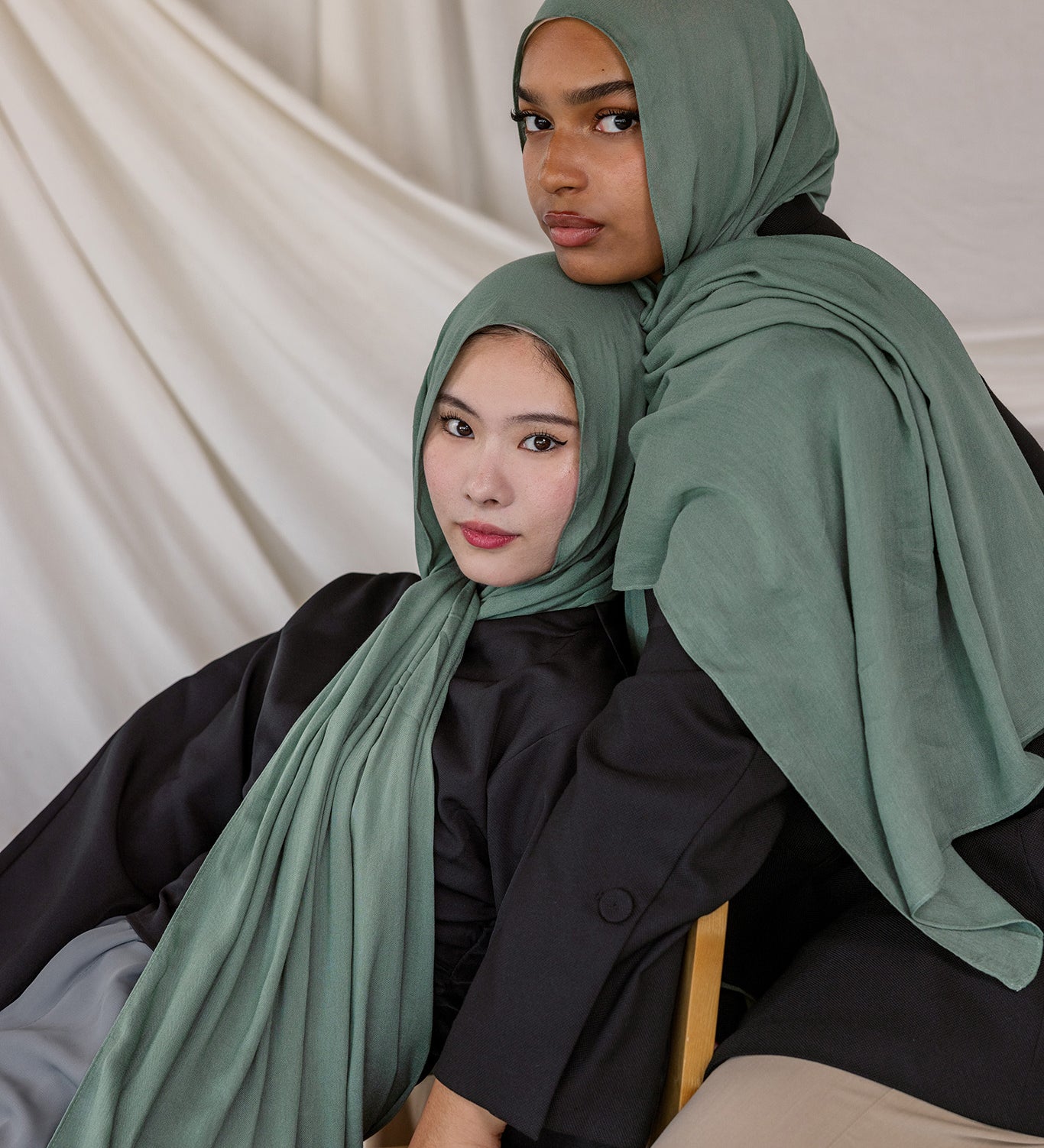 Two women wearing Pine Green hijabs sitting on a white draped background