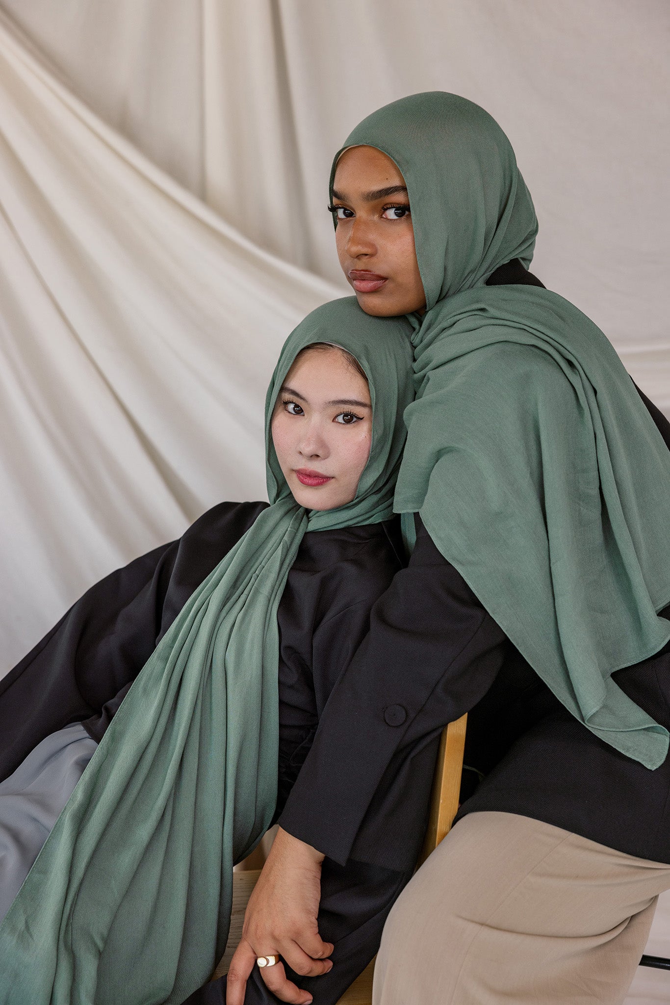 Two women wearing Pine Green hijabs sitting on a white draped background