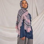Woman wearing a plaid hijab with pink lace over a blue top and gray pants against a beige curtain background