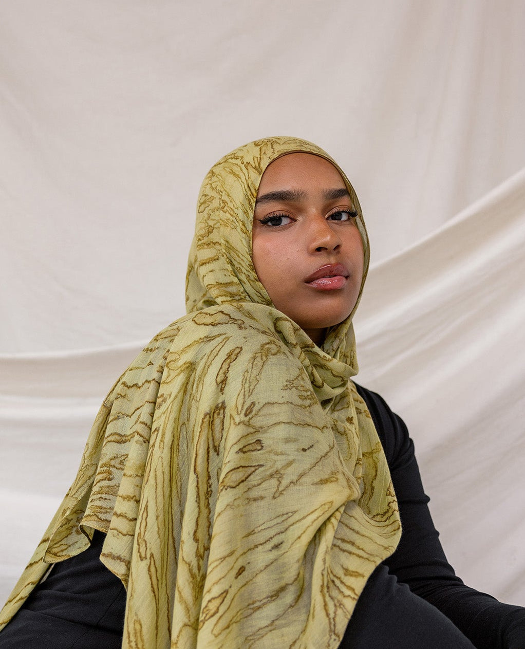 Model wearing a olive printed dune hijab and black outfit against a white draped background