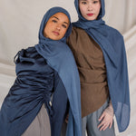 Two women wearing blue hijabs and gray pants against a plain background