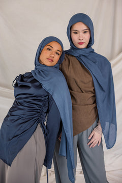 Two women wearing blue hijabs and gray pants against a plain background