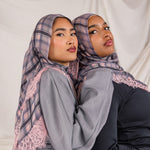 Two women wearing patterned pink plaid hijabs and gray tops against a plain background