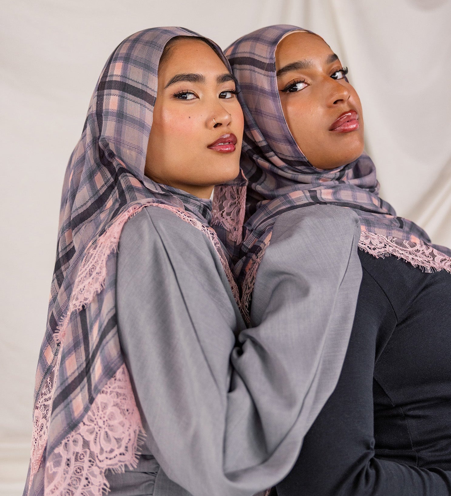 Two women wearing patterned pink plaid hijabs and gray tops against a plain background