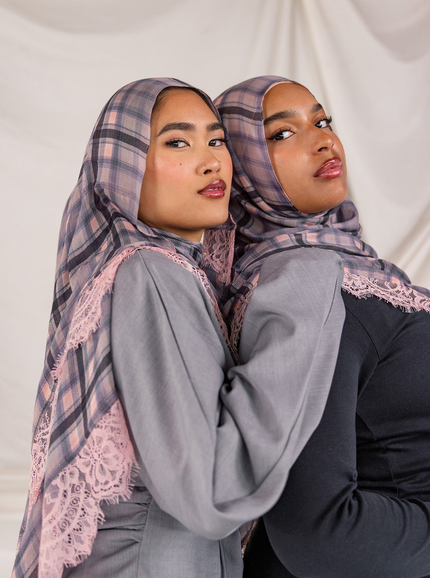 Two women wearing patterned pink plaid hijabs and gray tops against a plain background