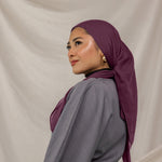 Woman wearing a purple red hijab and gray outfit against a beige background