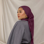 Woman wearing a purple red undertoned Fig Latte headscarf and gray outfit against a beige background