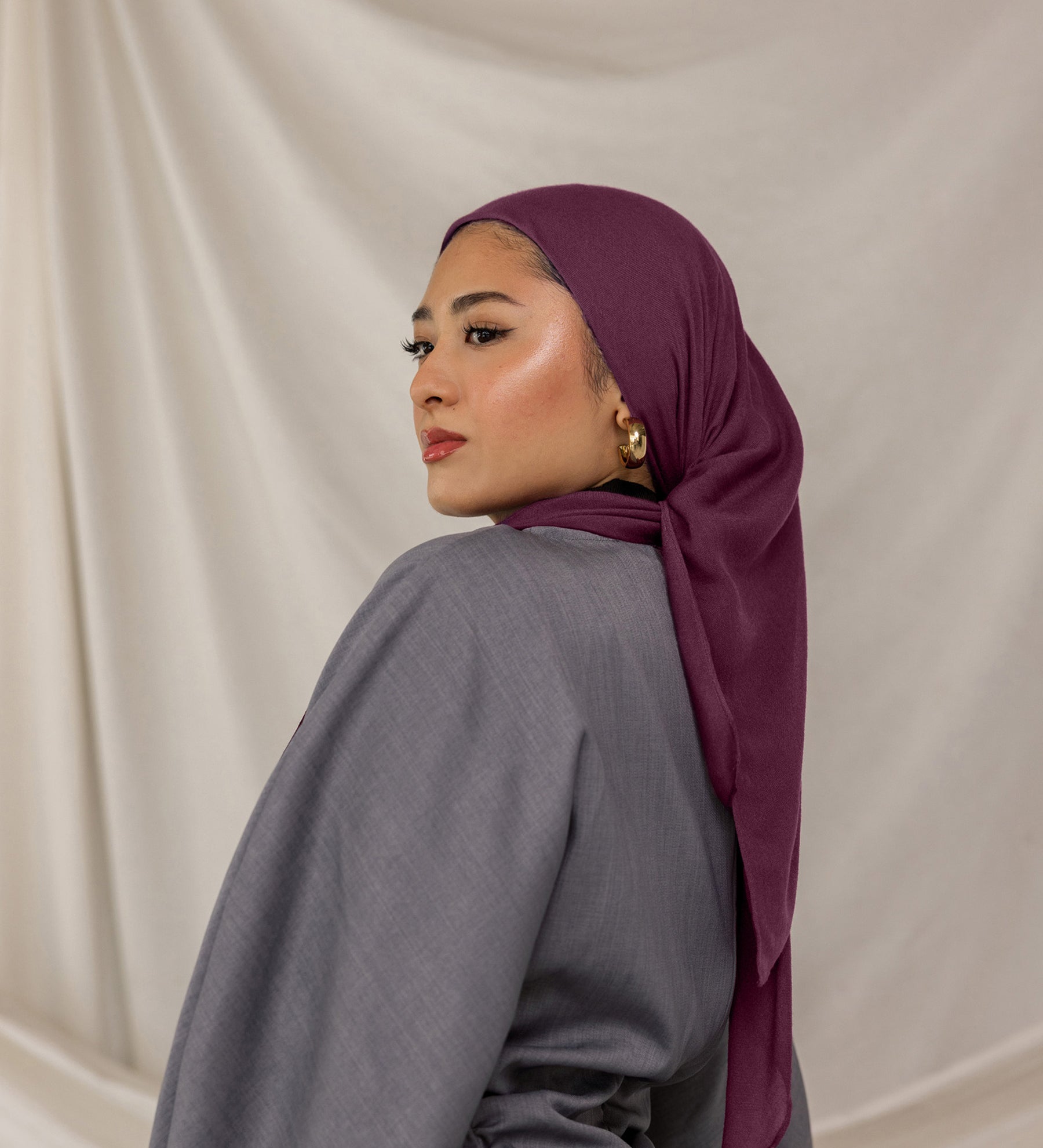 Woman wearing a purple red undertoned Fig Latte headscarf and gray outfit against a beige background