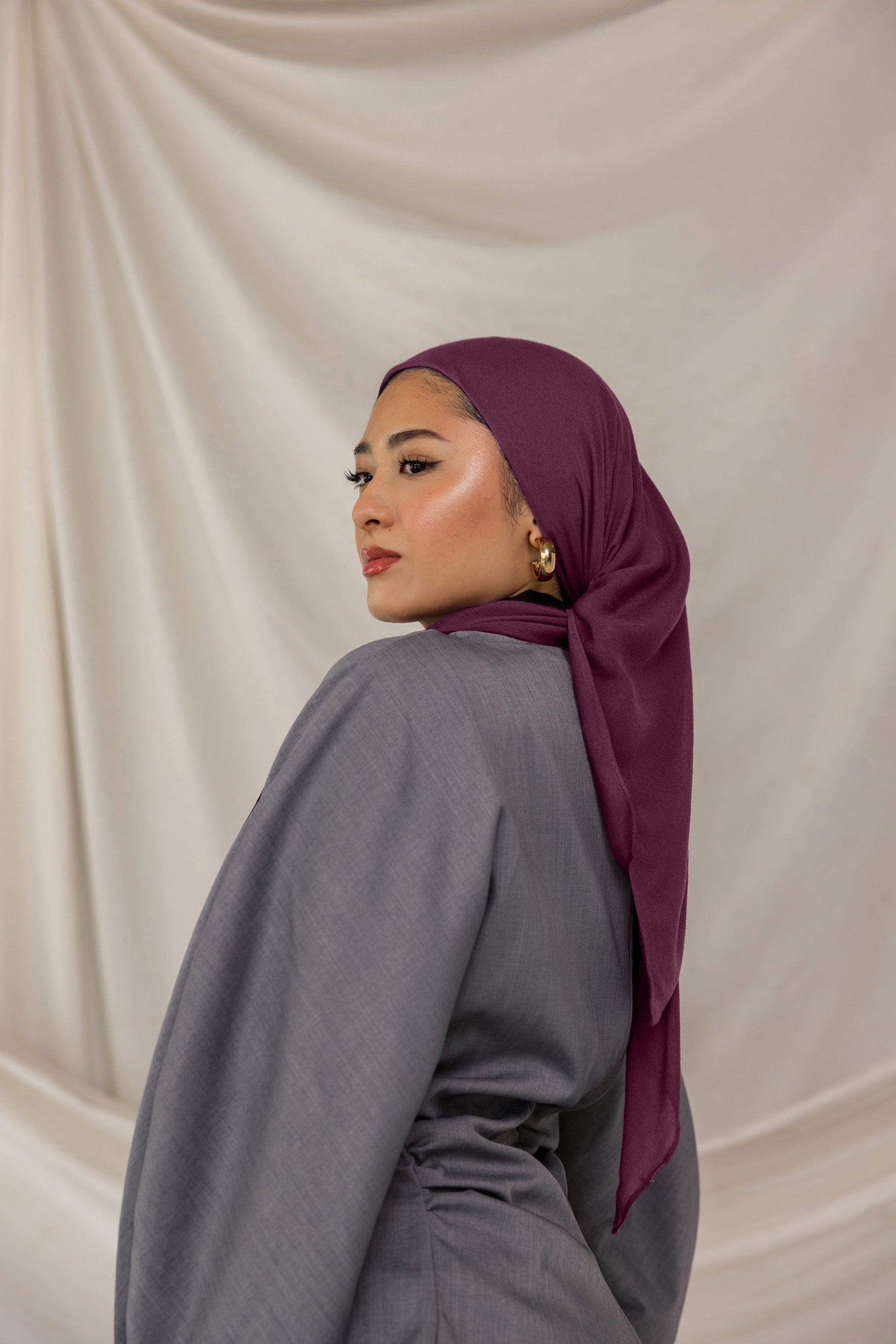 Woman wearing a purple red undertoned Fig Latte headscarf and gray outfit against a beige background