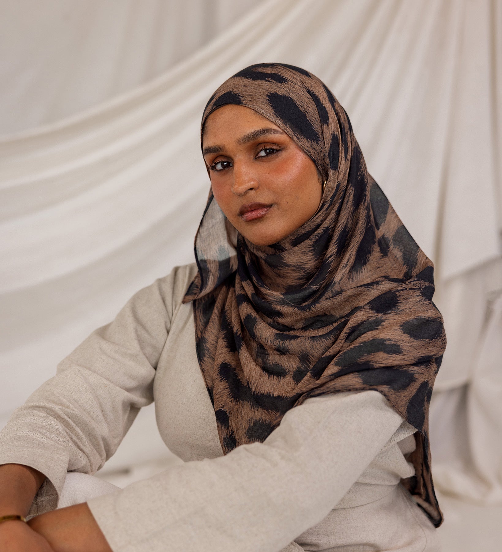 Model styled in a patterned leopard hijab sitting against a white draped background