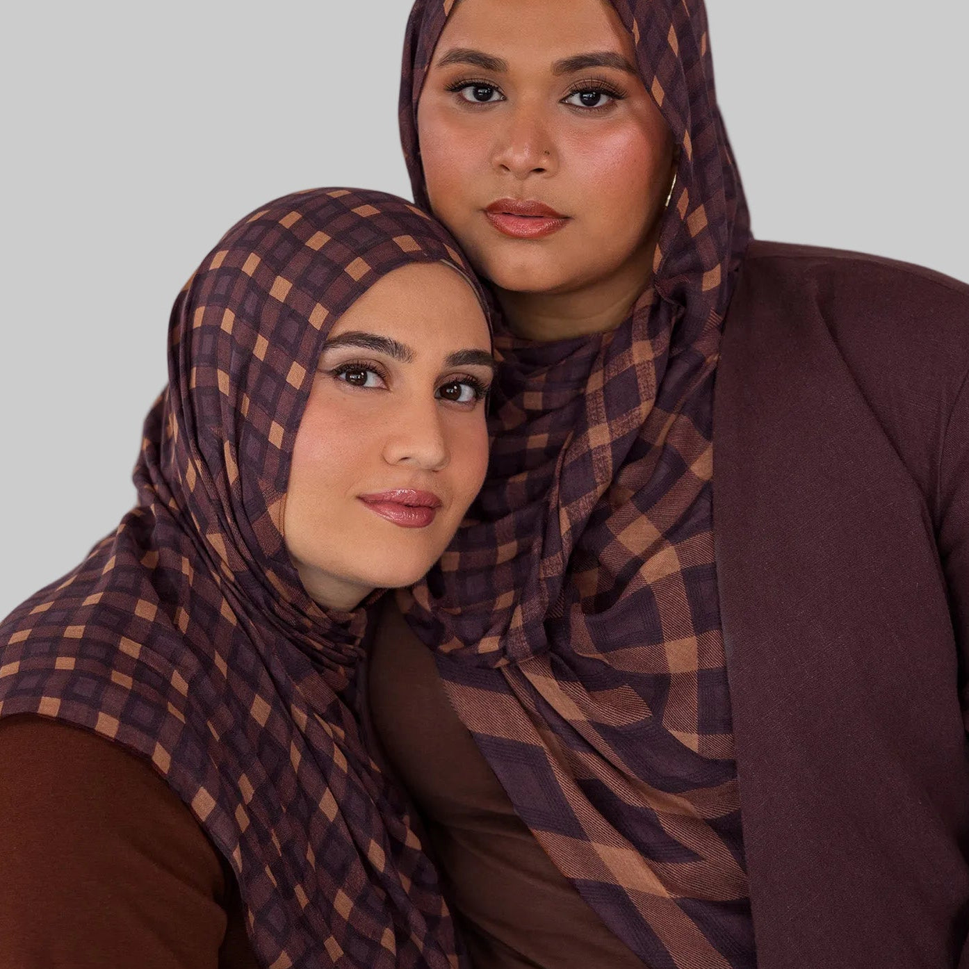 Two women wearing matching brown and purple checkered plaid print hijabs against a plain background