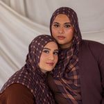Two women wearing matching brown and purple checkered plaid print hijabs against a plain background