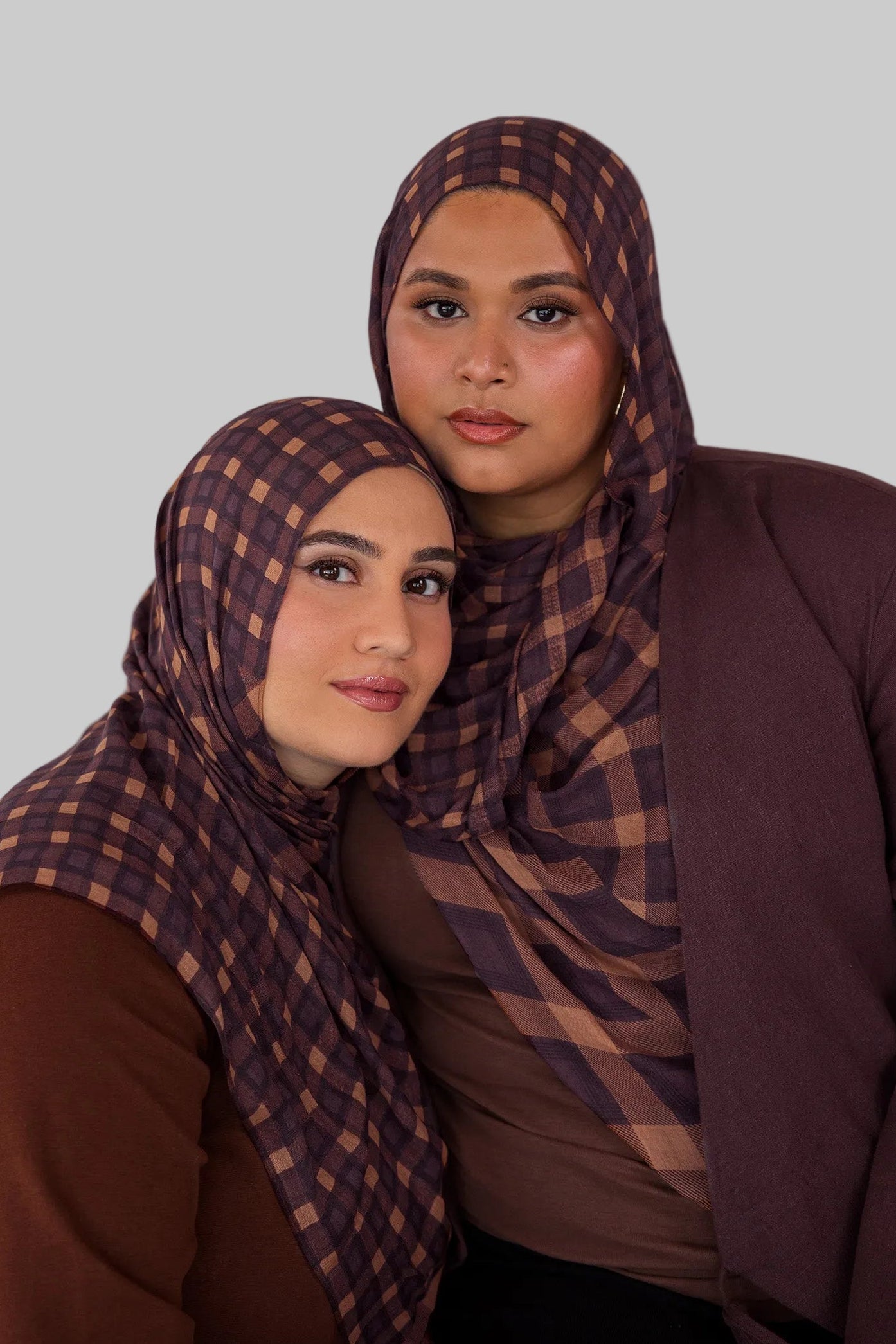Two women wearing matching brown and purple checkered plaid print hijabs against a plain background