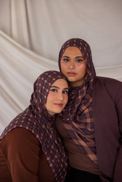 Two women wearing matching brown and purple checkered plaid print hijabs against a plain background