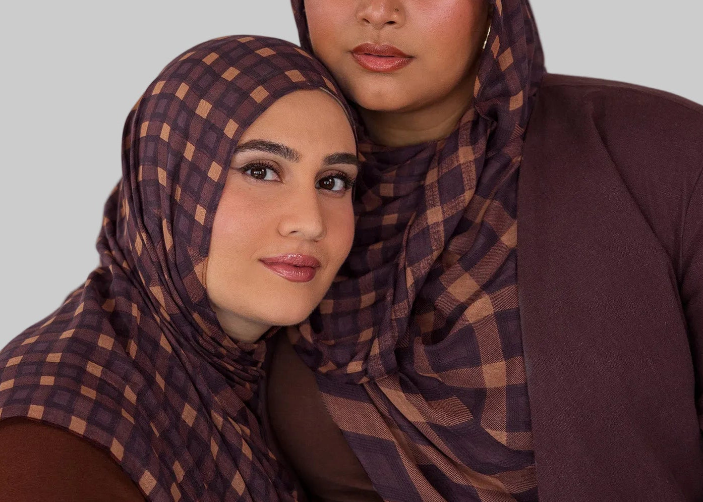 Two women wearing matching brown and purple checkered plaid print hijabs against a plain background