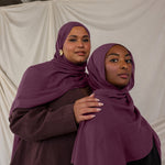 Two women wearing purple red toned hijabs standing close to each other against a plain background.