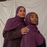 Two women wearing purple red toned hijabs standing close to each other against a plain background.