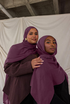 Two women wearing purple red toned hijabs standing close to each other against a plain background.