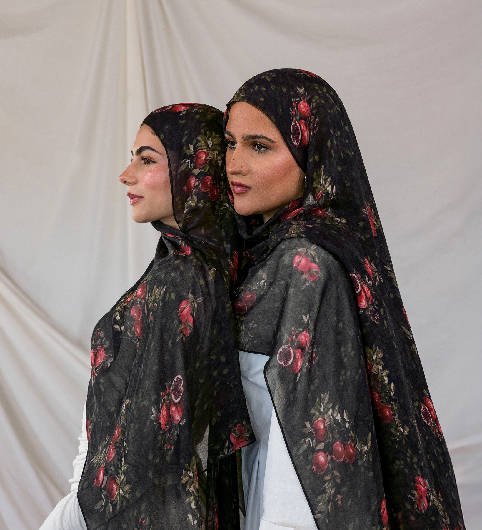 Two women wearing black pomegranate printed hijabs against a plain background