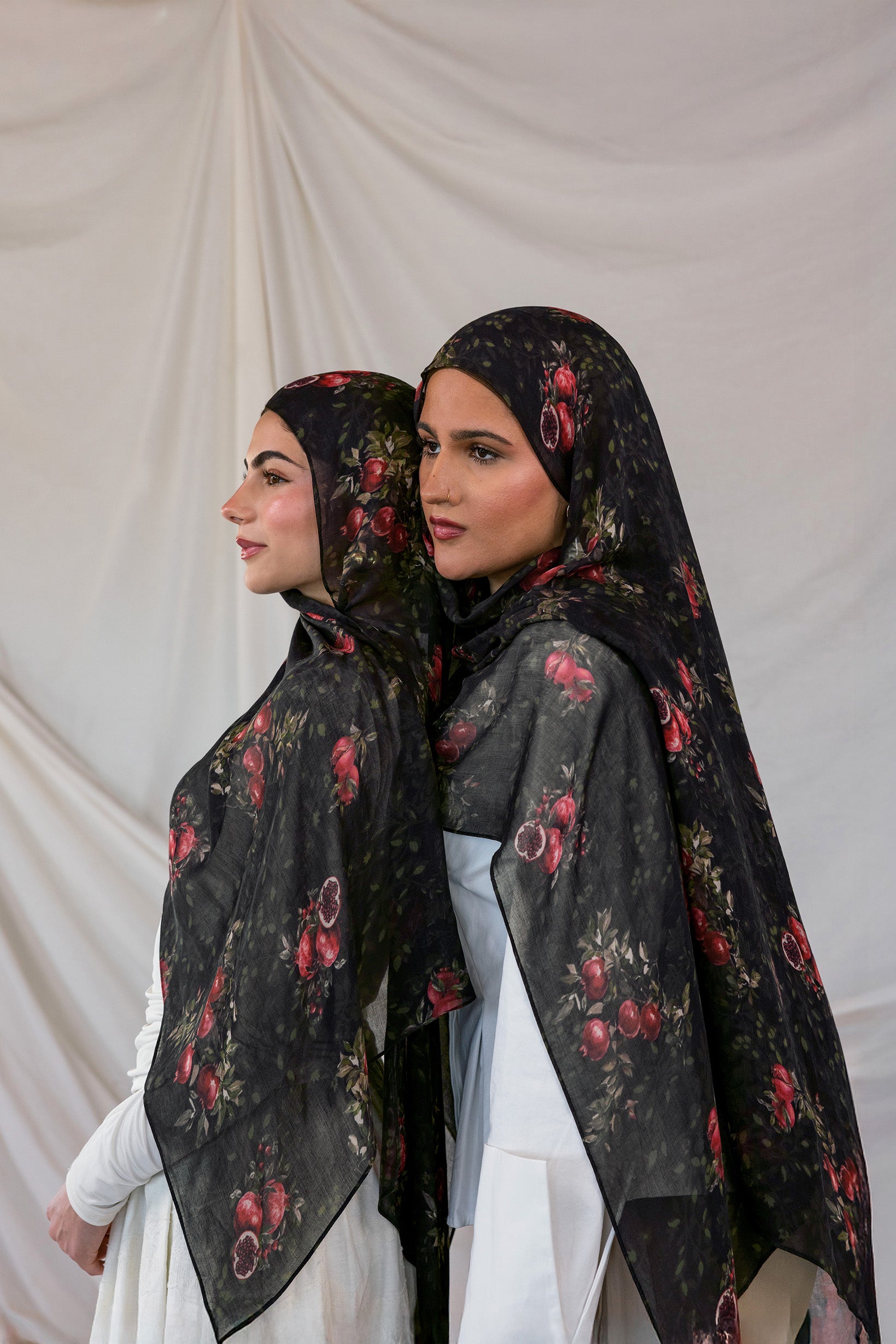 Two women wearing black pomegranate printed hijabs against a plain background