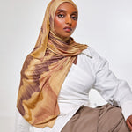 A woman wearing a beige and brown patterned hijab, a white button-up shirt, and taupe trousers, posing against a light gray studio background.