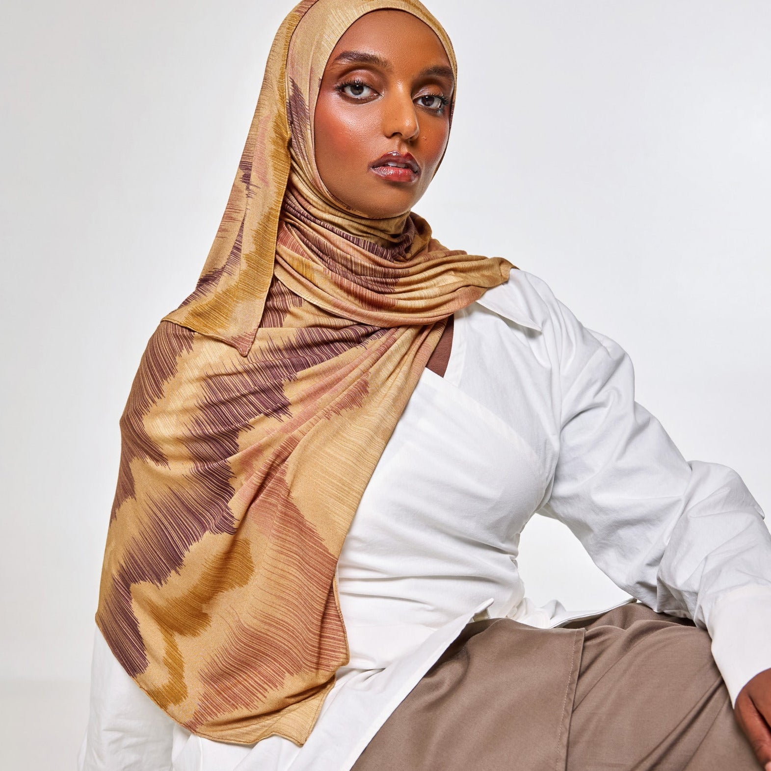A woman wearing a beige and brown patterned hijab, a white button-up shirt, and taupe trousers, posing against a light gray studio background.