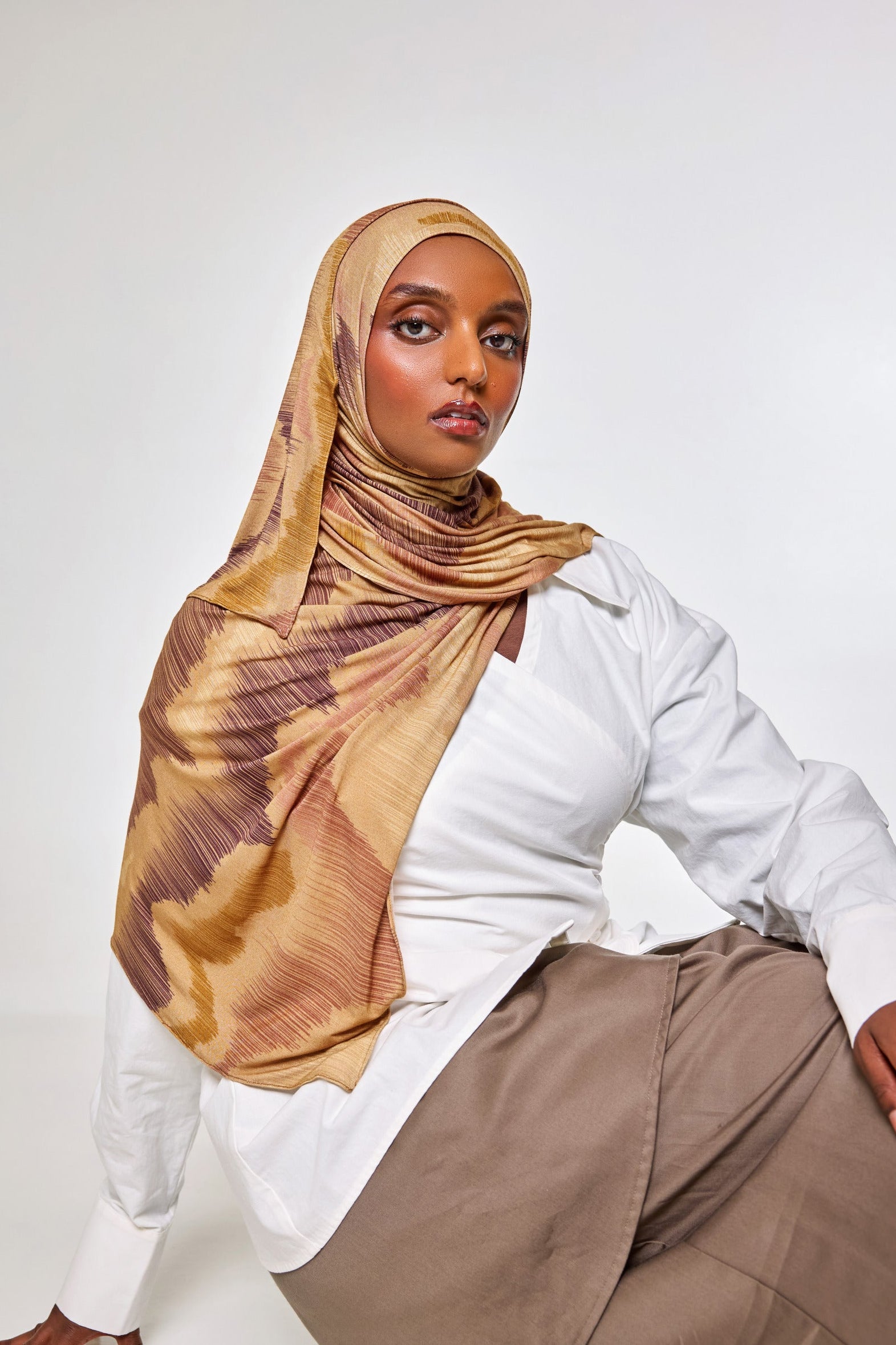 A woman wearing a beige and brown patterned hijab, a white button-up shirt, and taupe trousers, posing against a light gray studio background.