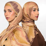 Two women shown from the back wearing matching patterned hijabs in beige, tan, and brown tones, with one in a beige jacket and the other in a brown top, against a light gray studio background.