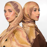 Two women shown from the back wearing matching patterned hijabs in beige, tan, and brown tones, with one in a beige jacket and the other in a brown top, against a light gray studio background.