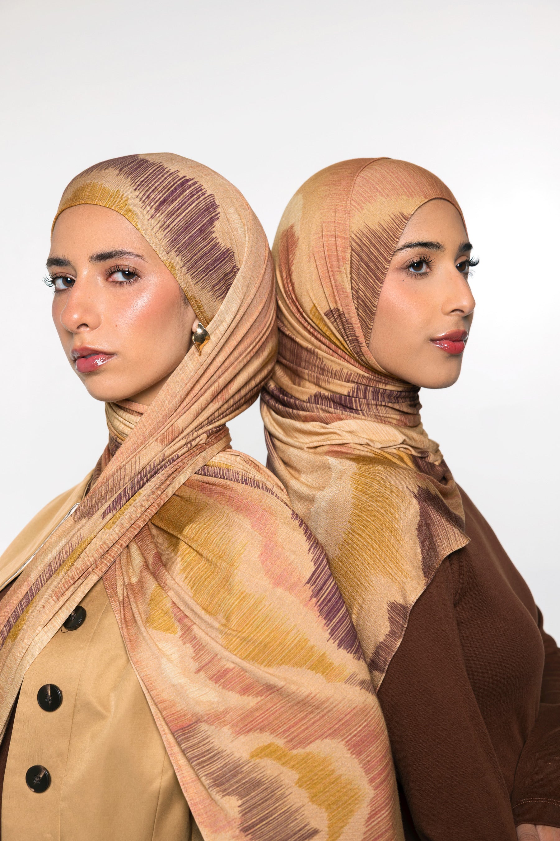Two women shown from the back wearing matching patterned hijabs in beige, tan, and brown tones, with one in a beige jacket and the other in a brown top, against a light gray studio background.