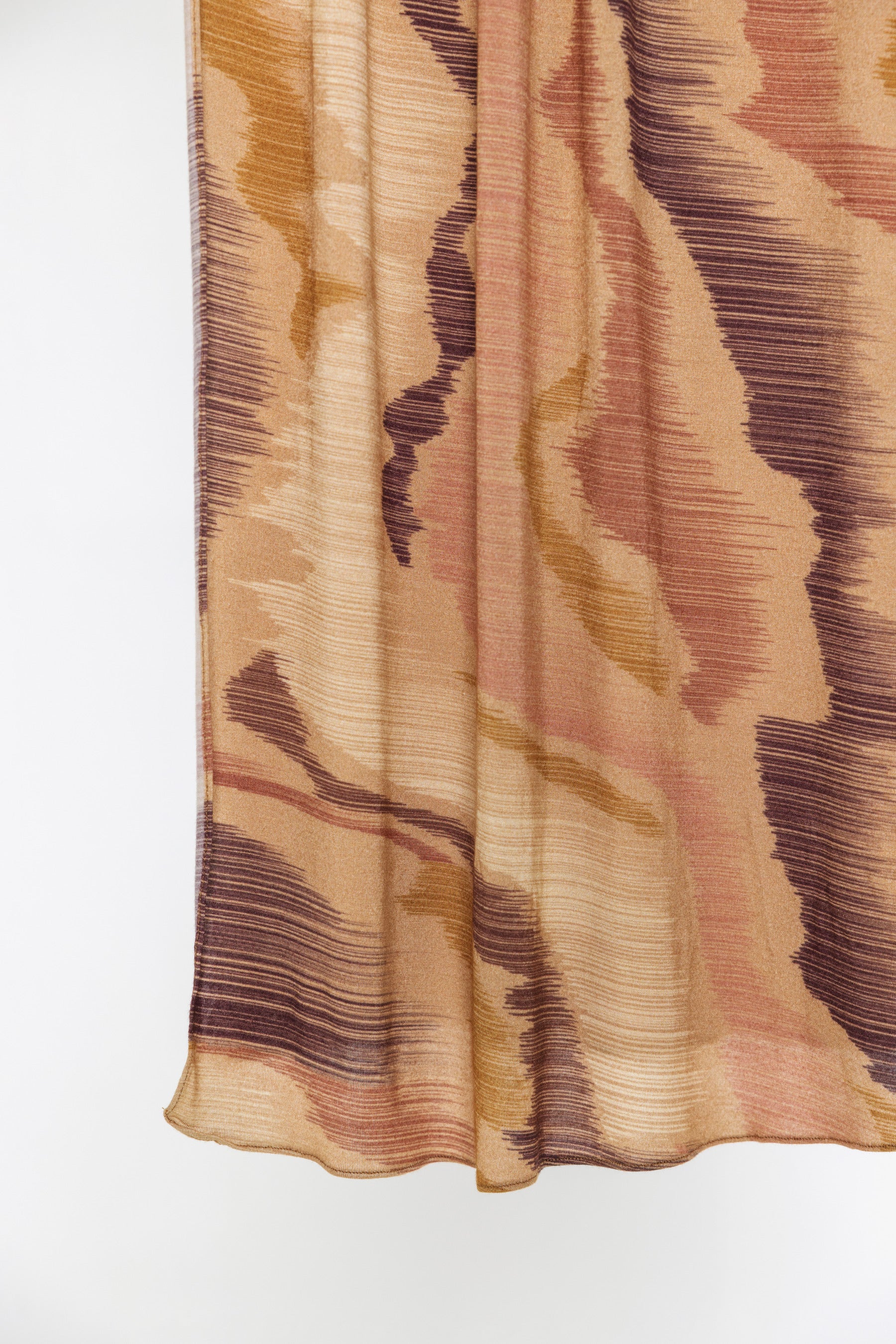 A close-up of a draped fabric with an abstract, textured pattern in shades of beige, brown, and muted pink on a plain white background.