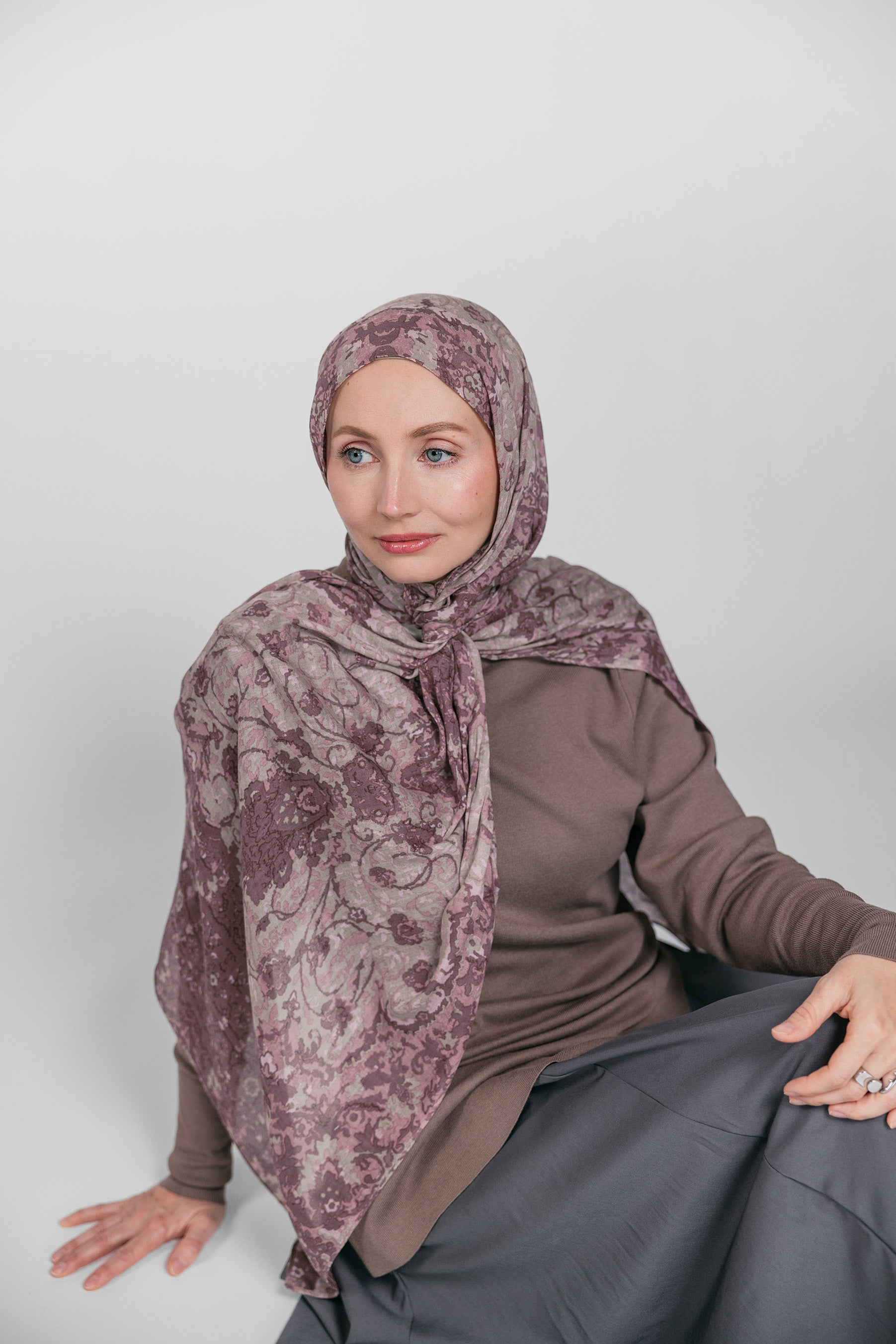 Woman wearing a patterned hijab, brown top, and gray skirt on a white background