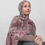 Woman wearing a mauve patterned hijab sitting against a plain background