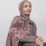 Woman wearing a mauve patterned hijab sitting against a plain background