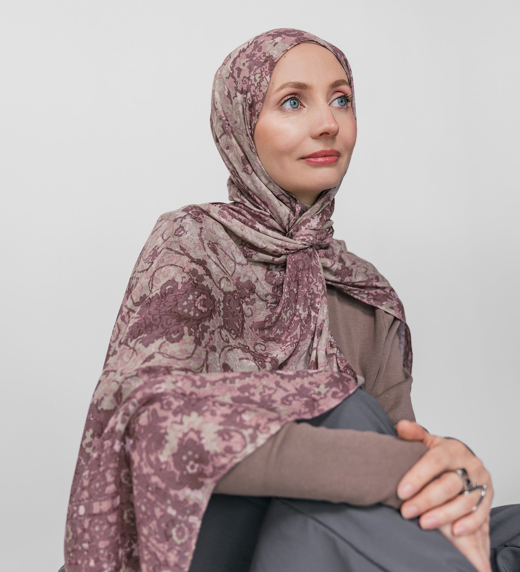 Woman wearing a mauve patterned hijab sitting against a plain background