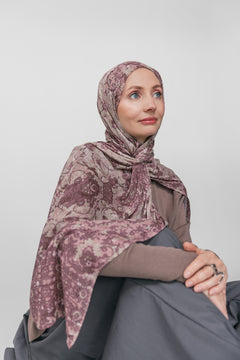 Woman wearing a mauve patterned hijab sitting against a plain background