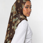 A woman wearing a brown hijab with a muted green and beige abstract floral pattern, paired with a white long-sleeve top, photographed against a plain light gray background.