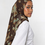 A woman wearing a brown hijab with a muted green and beige abstract floral pattern, paired with a white long-sleeve top, photographed against a plain light gray background.