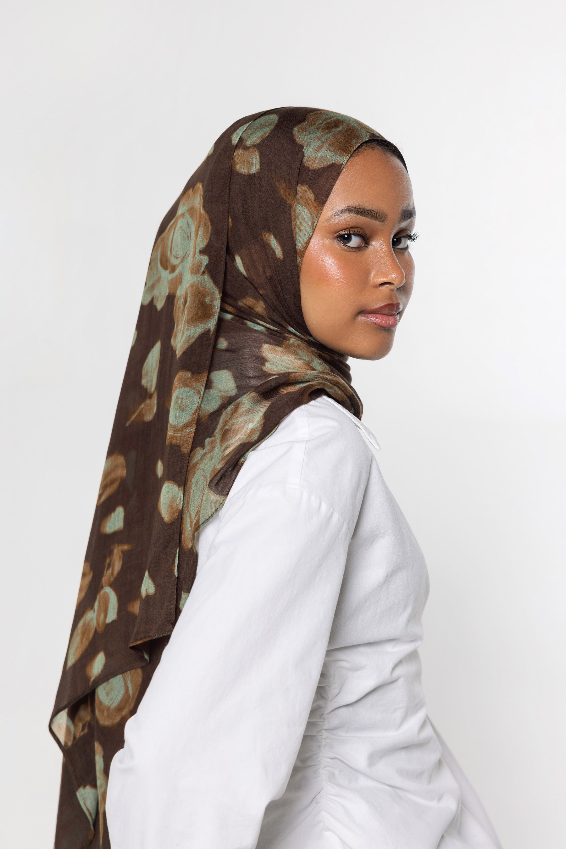 A woman wearing a brown hijab with a muted green and beige abstract floral pattern, paired with a white long-sleeve top, photographed against a plain light gray background.