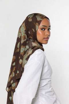 A woman wearing a brown hijab with a muted green and beige abstract floral pattern, paired with a white long-sleeve top, photographed against a plain light gray background.