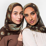 Two women wearing matching brown hijabs with a muted abstract pattern in beige and olive green, posed against a light gray studio background.