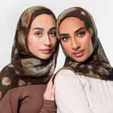 Two women wearing matching brown hijabs with a muted abstract pattern in beige and olive green, posed against a light gray studio background.