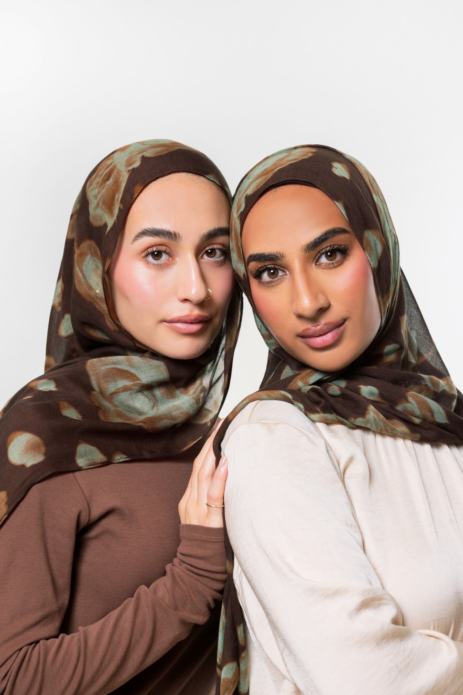 Two women wearing matching brown hijabs with a muted abstract pattern in beige and olive green, posed against a light gray studio background.