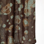 A close-up of a sheer brown fabric with an abstract floral pattern in muted beige and pale blue-green, hanging against a white background.