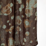 A close-up of a sheer brown fabric with an abstract floral pattern in muted beige and pale blue-green, hanging against a white background.