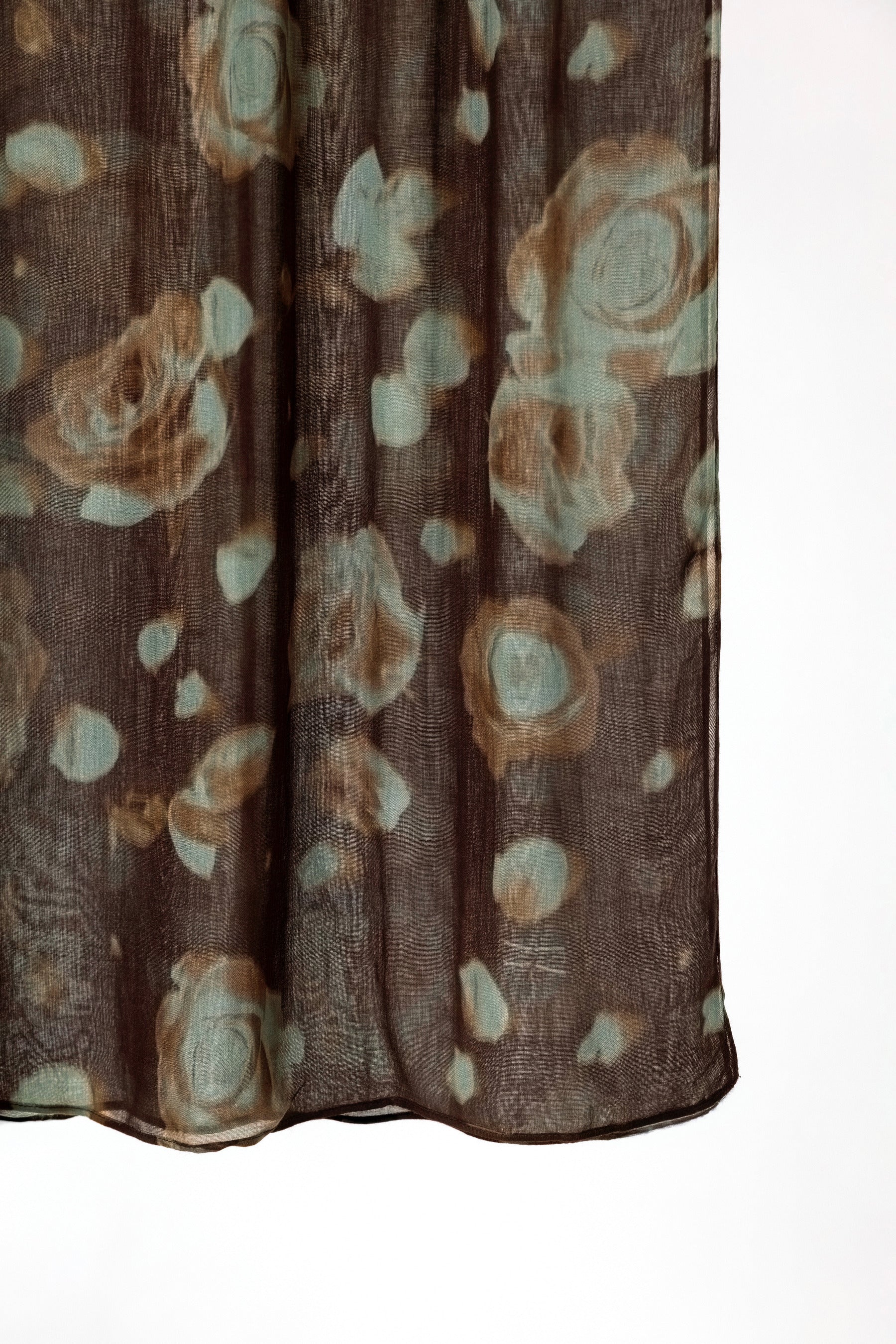 A close-up of a sheer brown fabric with an abstract floral pattern in muted beige and pale blue-green, hanging against a white background.